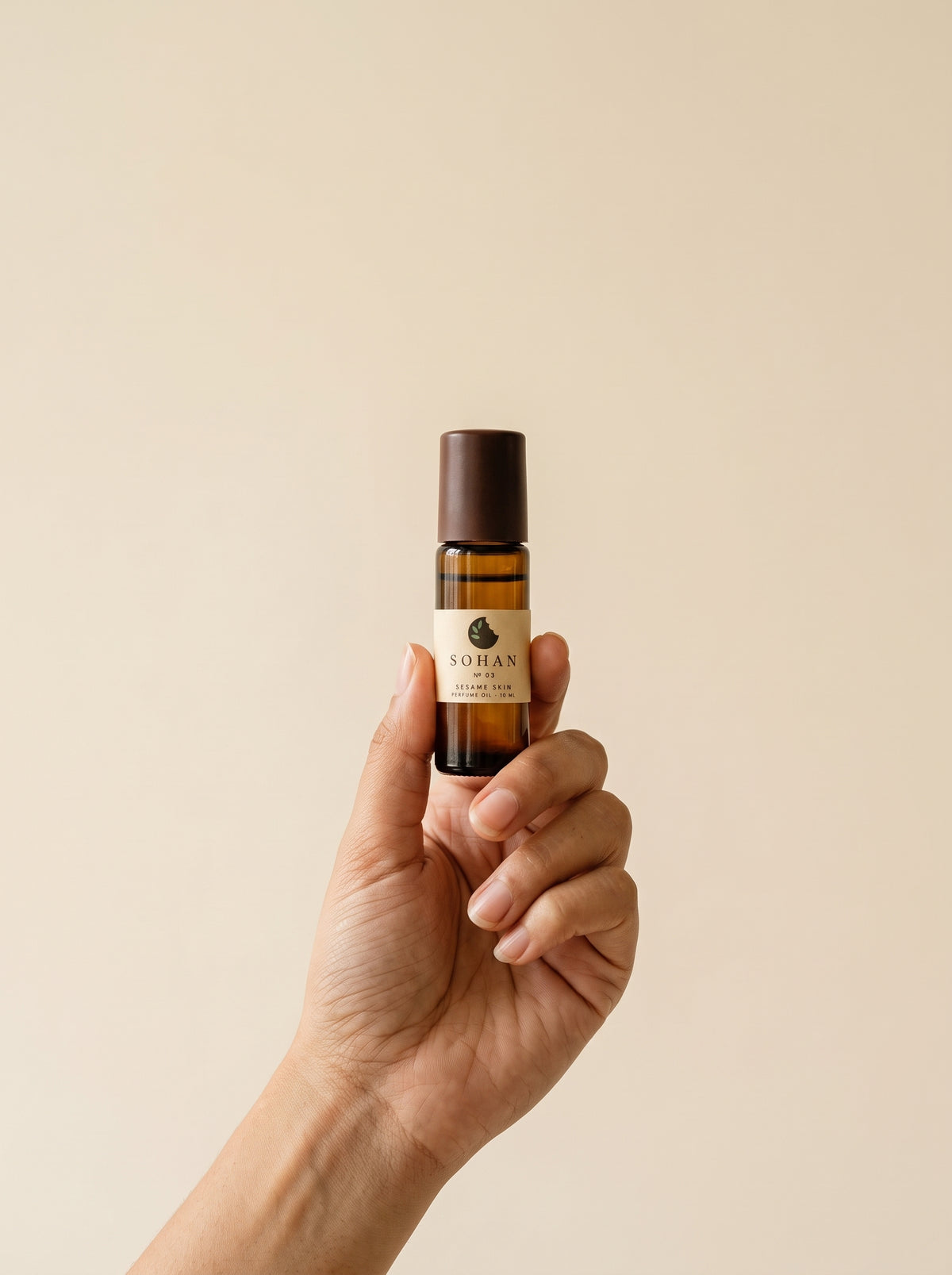 A hand holding a small amber glass roll-on bottle with a dark brown cap and a cream label that reads 'SOILAN'. The background is a solid, warm beige color.